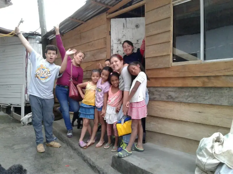 Una voluntaria y un niño con los brazos arriba, junto a niñas, frente a una casita de madera