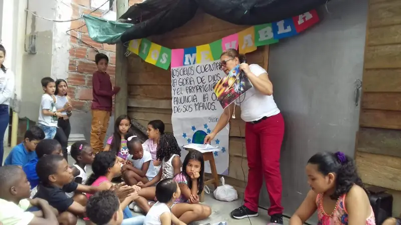 Una voluntaria mostrando un cartel a niños sentados en el piso, junto a un rancho de madera