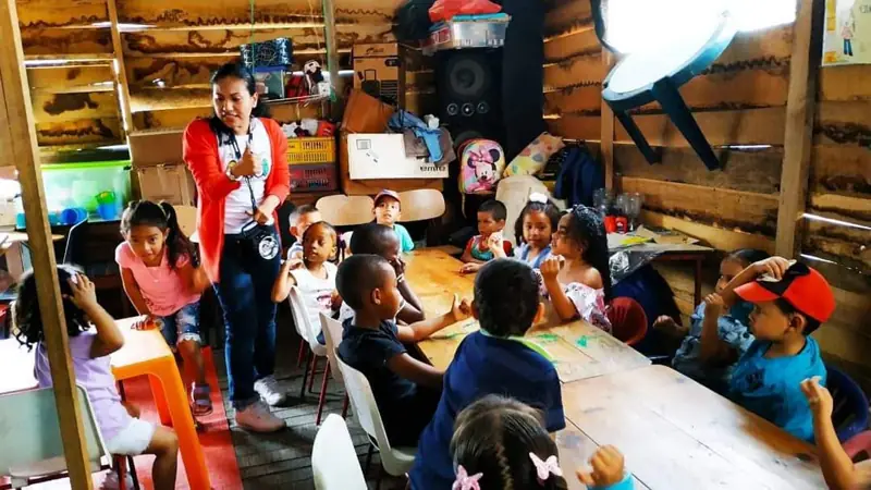 Una voluntaria de pie entre las mesas de los niños en el salón de madera