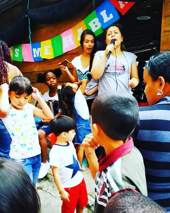 Una voluntaria cantando con micrófono frente a niños, bajo un banderín de colores