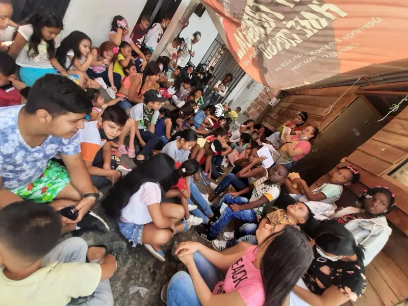 Muchos niños sentados en el piso de un pasillo largo con paredes de madera