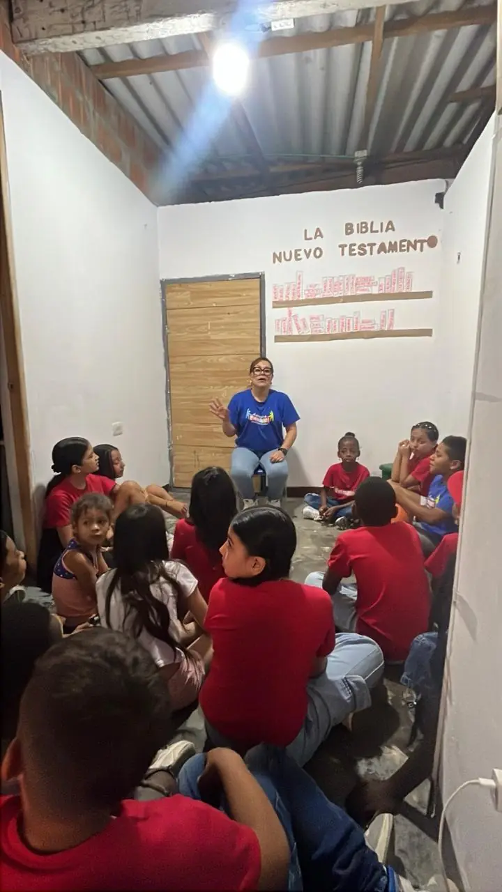 Niños con camisetas rojas escuchando a una voluntaria en un salón con un mural de la Biblia