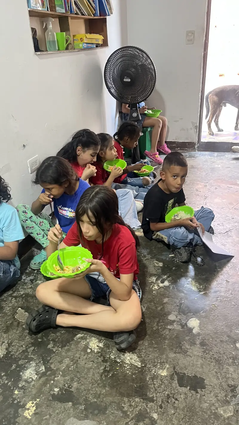 Niños y niñas almorzando sentados en el piso del comedor, con platos verdes