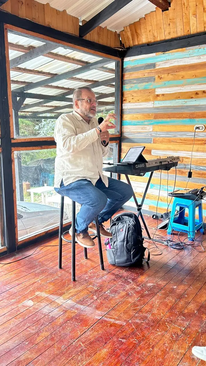 A teacher seated on a stool speaking into a microphone, next to a keyboard, in a wooden room